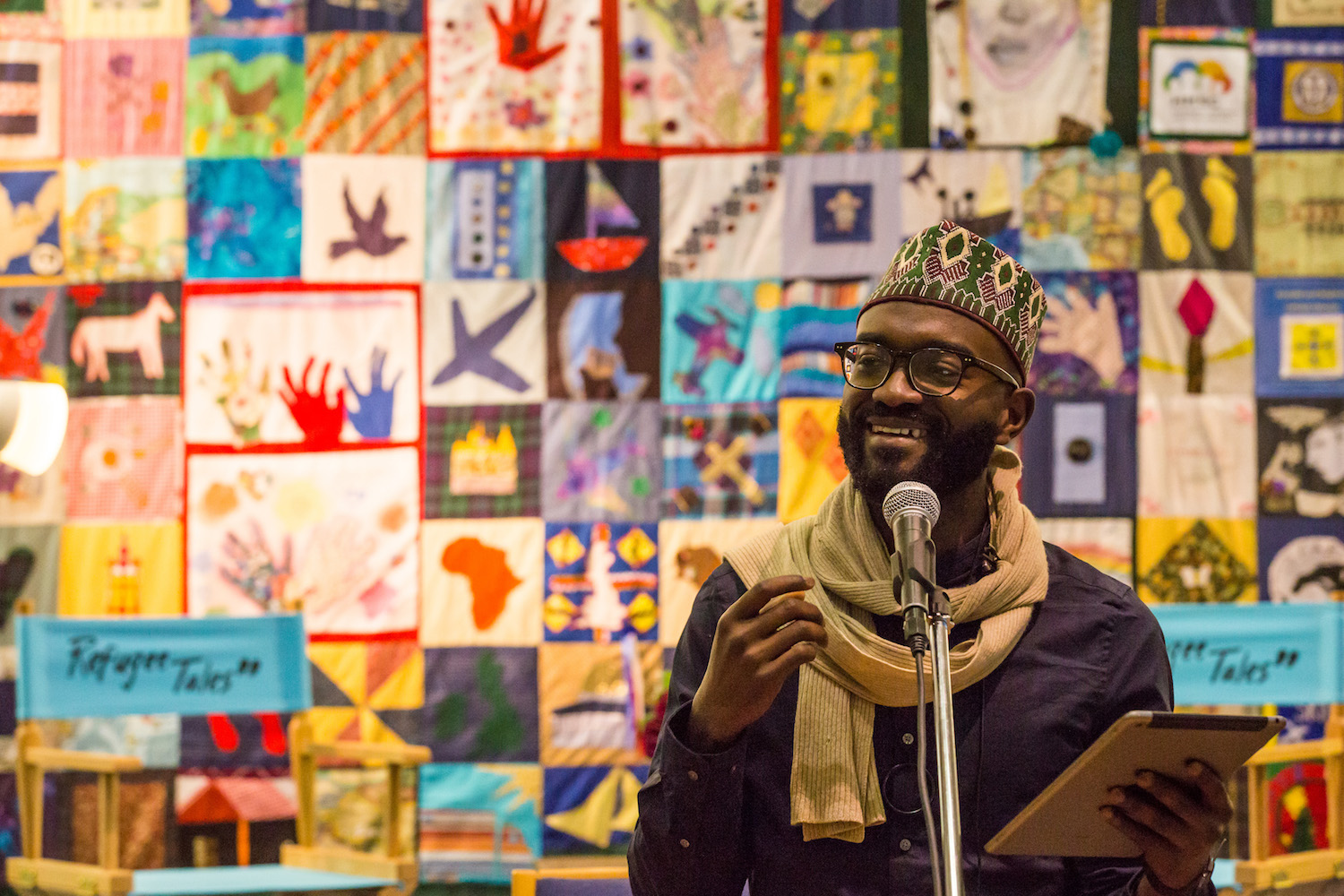 Poet, playwright and writer Inua Ellams reading for Refugee Tales in front of quilt made by Gatwick Detainees Welfare Group Quilt presented by Andrea Luka Zimmerman in The London Open 2018. Courtesy Gatwick Detainees Welfare Group and Refugee Tales. Photo by Chris Orange