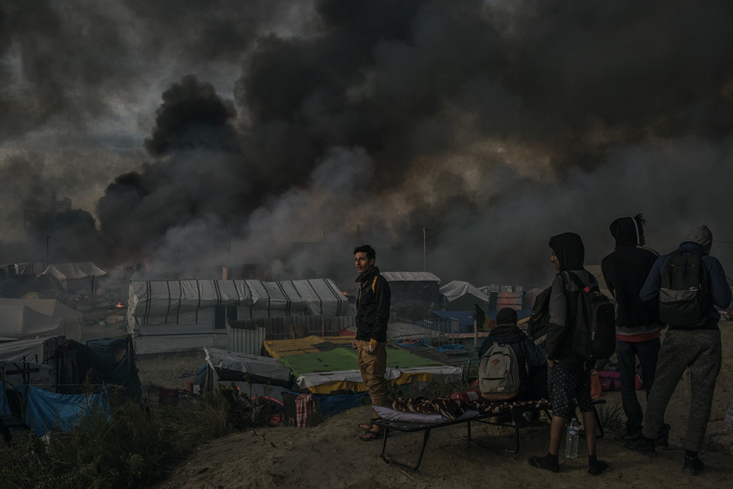 MAURICIO LIMARefugees watch a huge plume of smoke as dozens of res burn huts and makeshift shops at the camp called the â€œJungleâ€, in Calais, northern France October 26, 2016. Â© Mauricio Lima for The New York Times